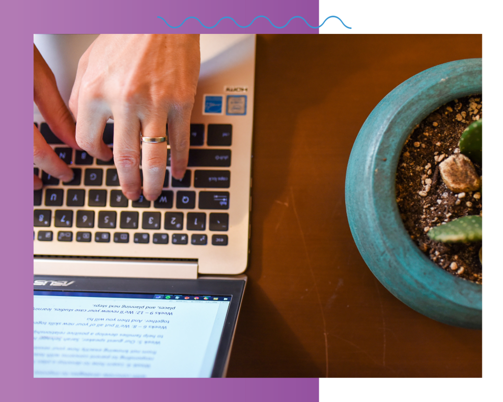 Two light toned hands are typing on a computer. There is a cactus next to the computer on a brown desk.